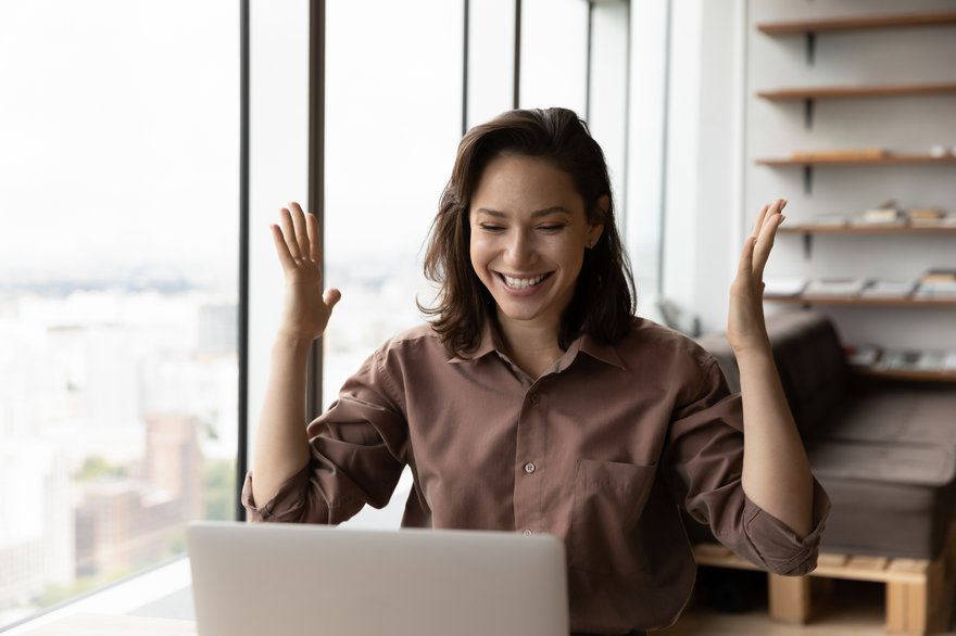 Smiling adult raises hands in celebration while seated at desk in front of laptop.