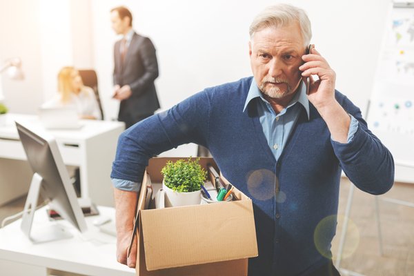Older man on a cell phone leaving an office with personal belongings in a box.