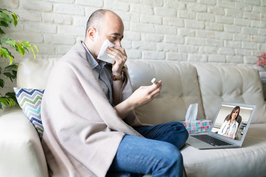 Sick person wrapped in blanket and blowing nose while attending telehealth appointment.