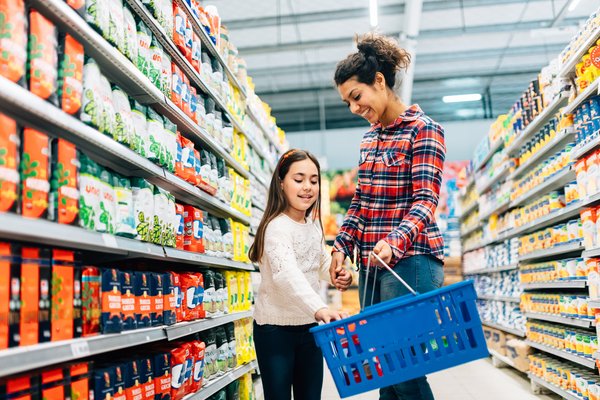 Parent and child browsing grocery aisle and placing items in blue shopping basket.