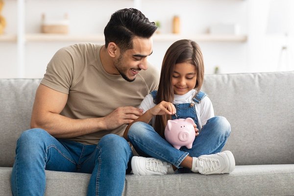 Child and adult sitting on a couch with child dropping coins into a piggy bank.