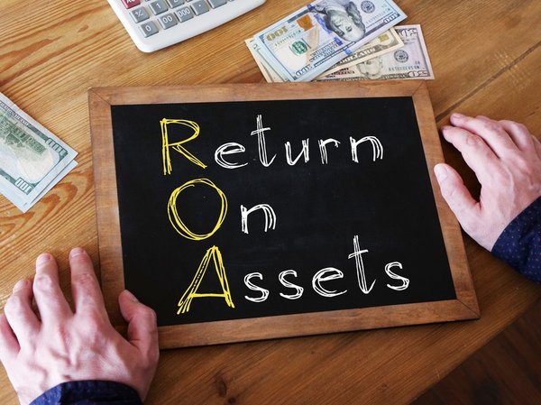 Hands next to a chalkboard with "Return on Assets" written on it and cash and a calculator next to the chalkboard