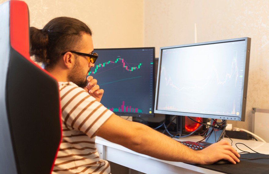 A man sitting at a desk with two computer monitors, one displaying a stock chart.