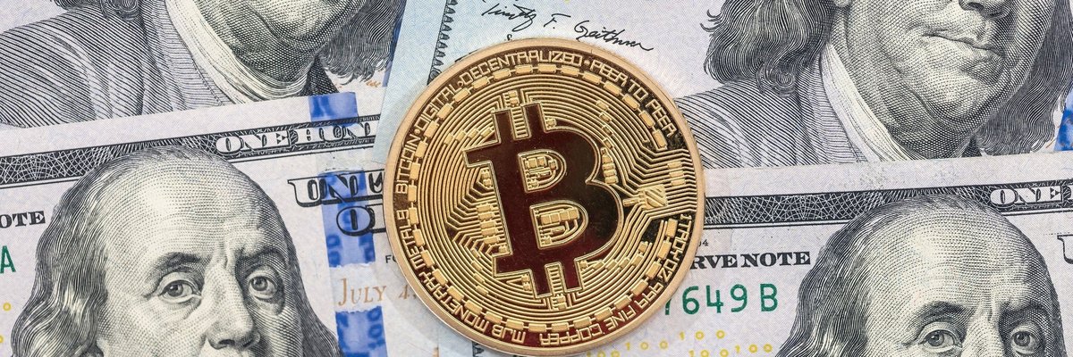 A physical gold coin marked with the Bitcoin B lying atop hundred dollar bills.