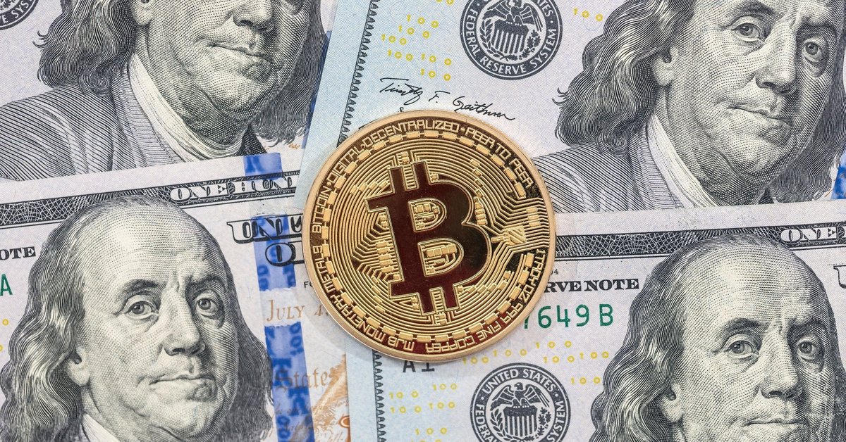 A physical gold coin marked with the Bitcoin B lying atop hundred dollar bills.