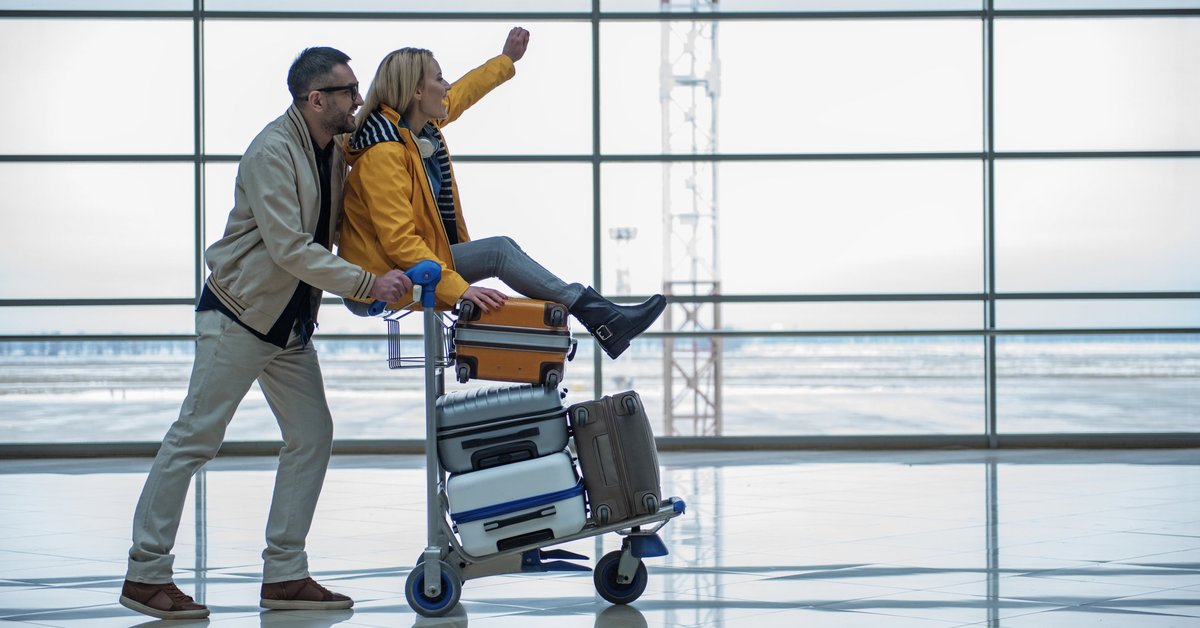 Excited woman riding luggage cart in airport with man pushing her.
