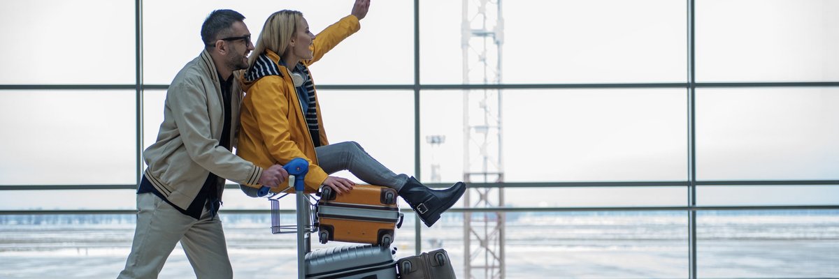 Excited woman riding luggage cart in airport with man pushing her.