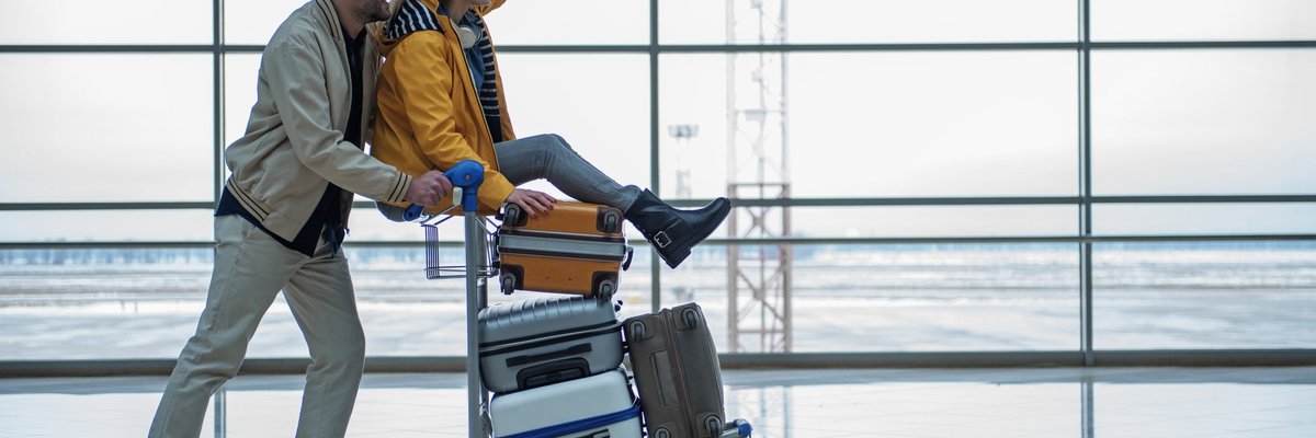 A man pushing a luggage cart through an airport with a cheering woman on top.
