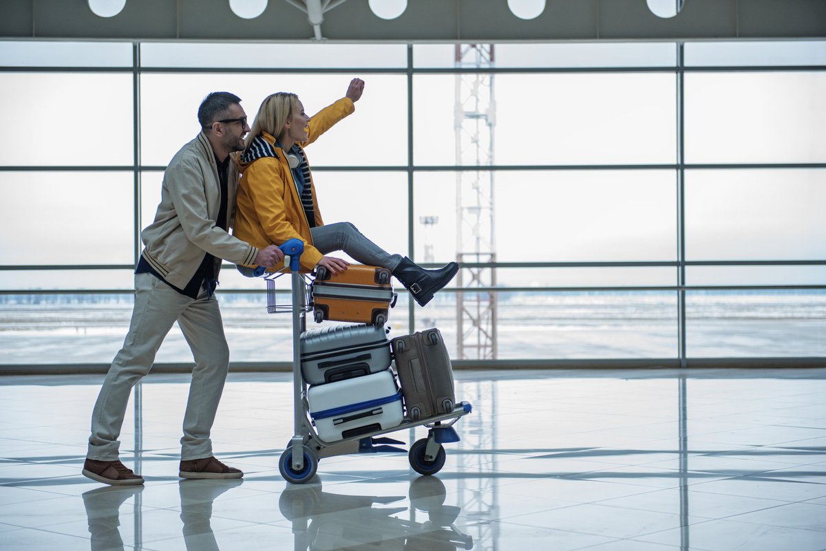 A man pushing a luggage cart through an airport with a cheering woman on top.