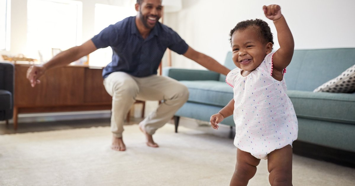 Baby fist-pumping and dad smiling with his arms open in the background