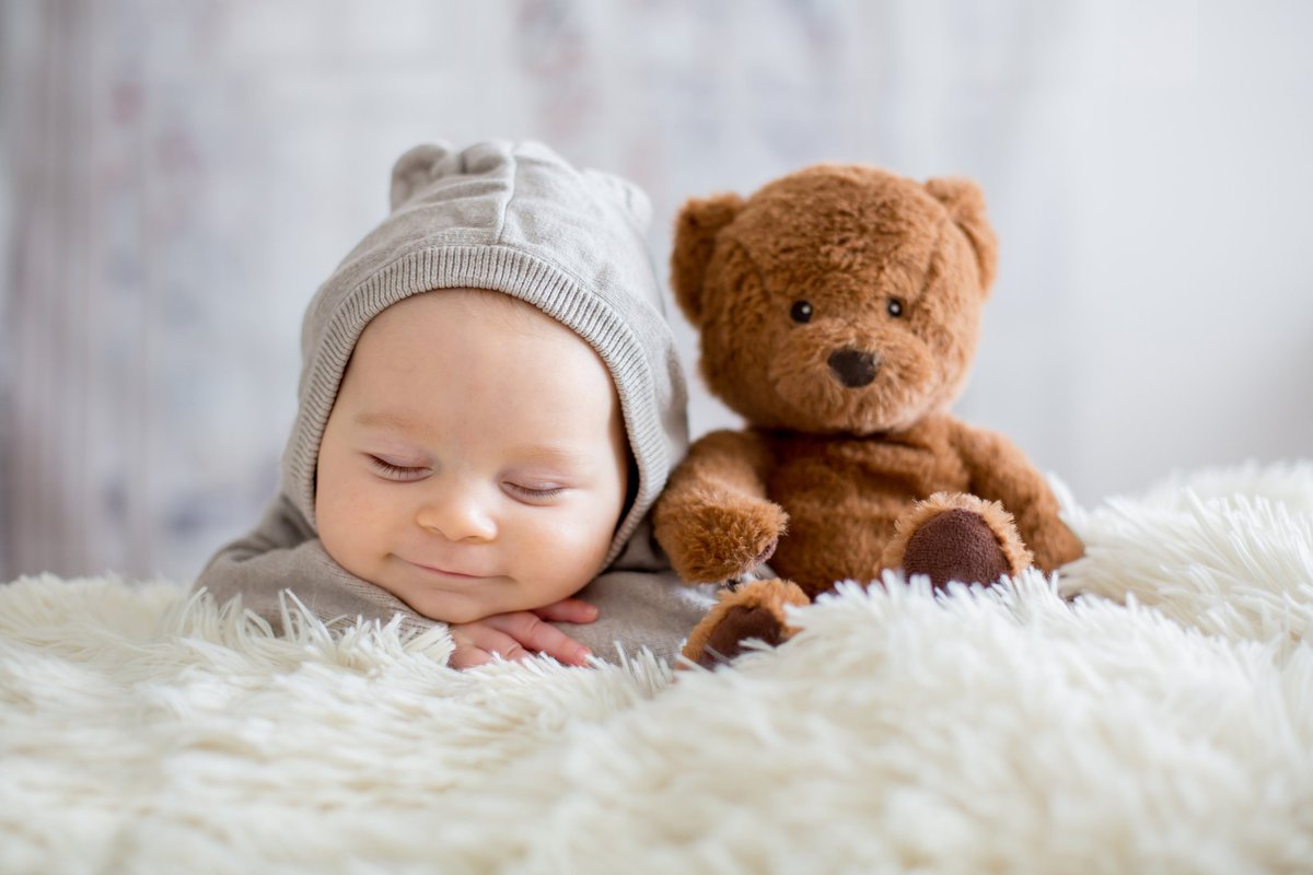 Sleepy, smiling baby in a knit hat next to a teddy bear on a fuzzy blanket.