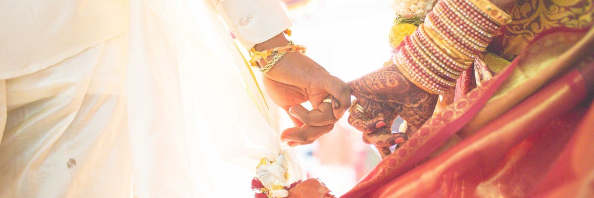 A bride with henna and bracelets on her arm holds hands with a groom.