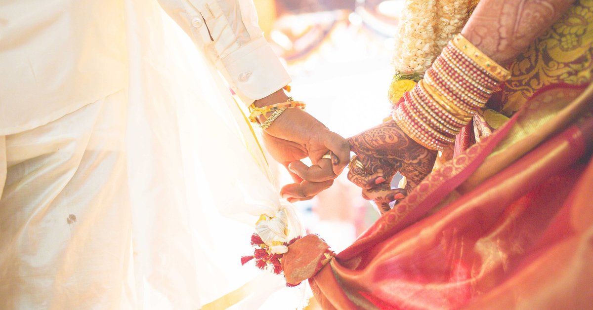 A bride with henna and bracelets on her arm holds hands with a groom.