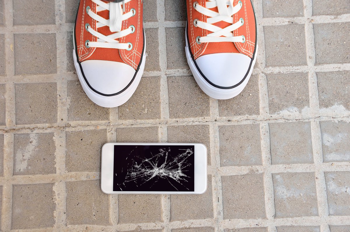 Someone standing next to a dropped cell phone with a broken screen.