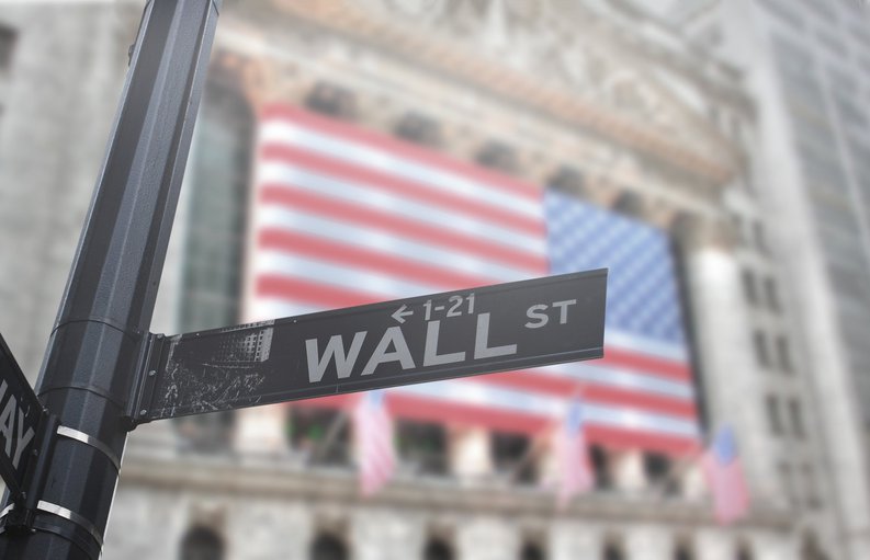 A street sign that says Wall St. with the American flag on a building in the background.