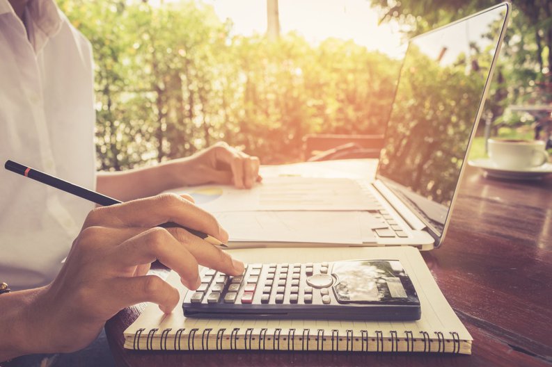 A person typing on a calculator while sitting in front of a laptop and notebook at a sunny cafe table.