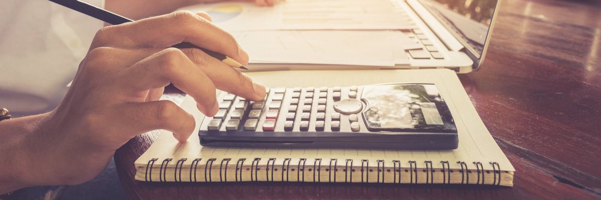 A person typing on a calculator while sitting in front of a laptop and notebook at a sunny cafe table.