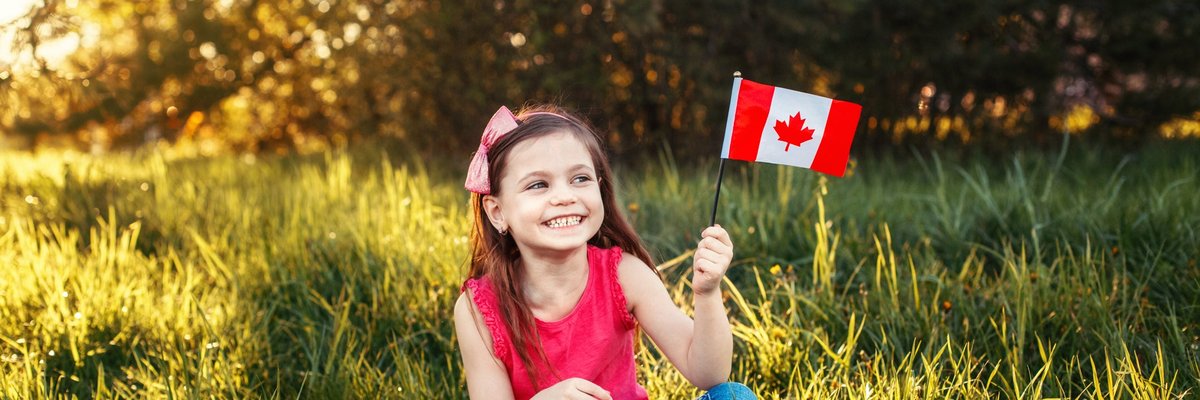 A child sits cross-legged in a meadow, smiling and holding a Canadian flag.