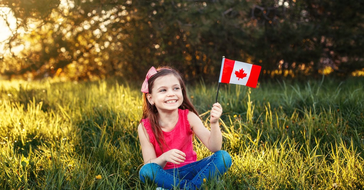 A child sits cross-legged in a meadow, smiling and holding a Canadian flag.