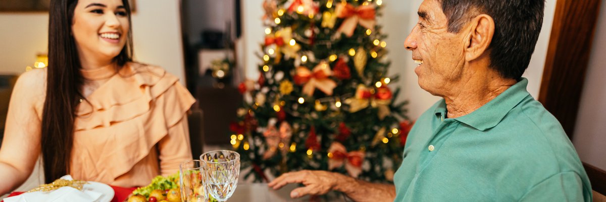 Family members sit at dinner table with decorated Christmas tree nearby.