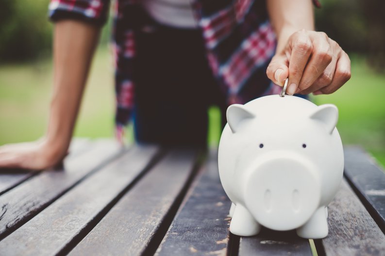 A person standing at a picnic table in a park and putting a coin in a piggy bank.