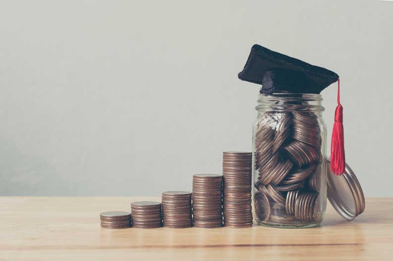 A pile of coins sit next to a jar full of coins and topped with a graduation cap.