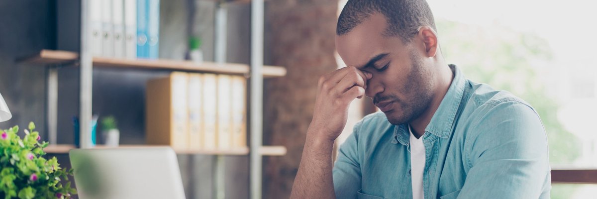 A distressed man sitting in front of a laptop with his hand on his forehead.