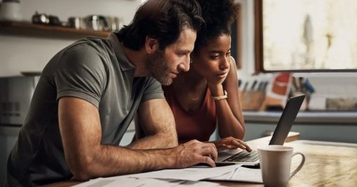 A man and woman sitting at their kitchen table with papers spread around them while researching something on a laptop.