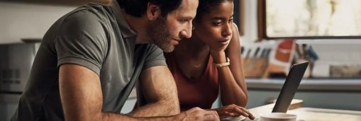 A man and woman sitting at their kitchen table with papers spread around them while researching something on a laptop.
