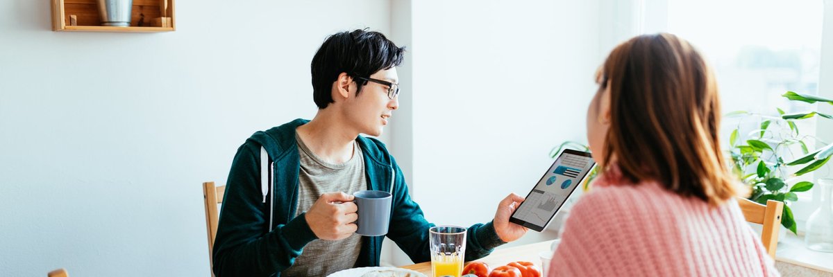 A couple eats breaksfast in their kitchen while the man looks at his tablet.