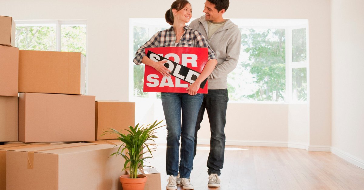 Couple holding sold real estate sign next to moving boxes.
