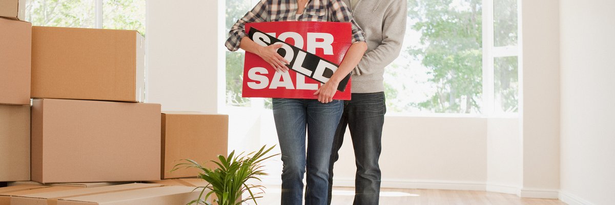 Couple holding sold real estate sign next to moving boxes.