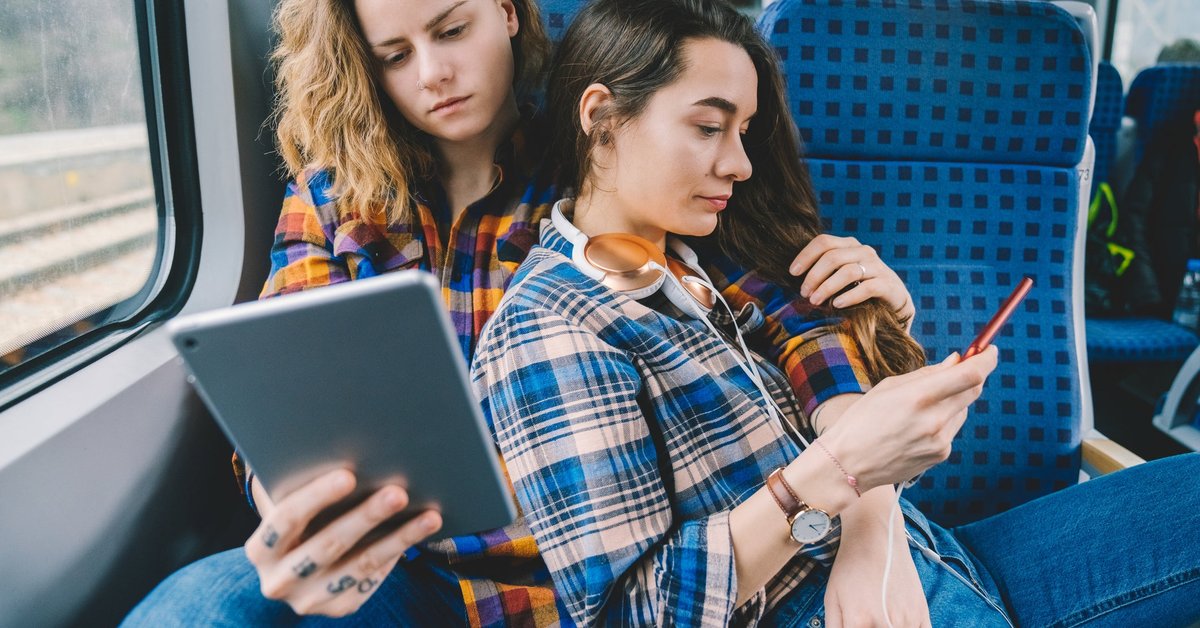 A couple sit together on a train. One looks at her tablet while the other looks at her phone.