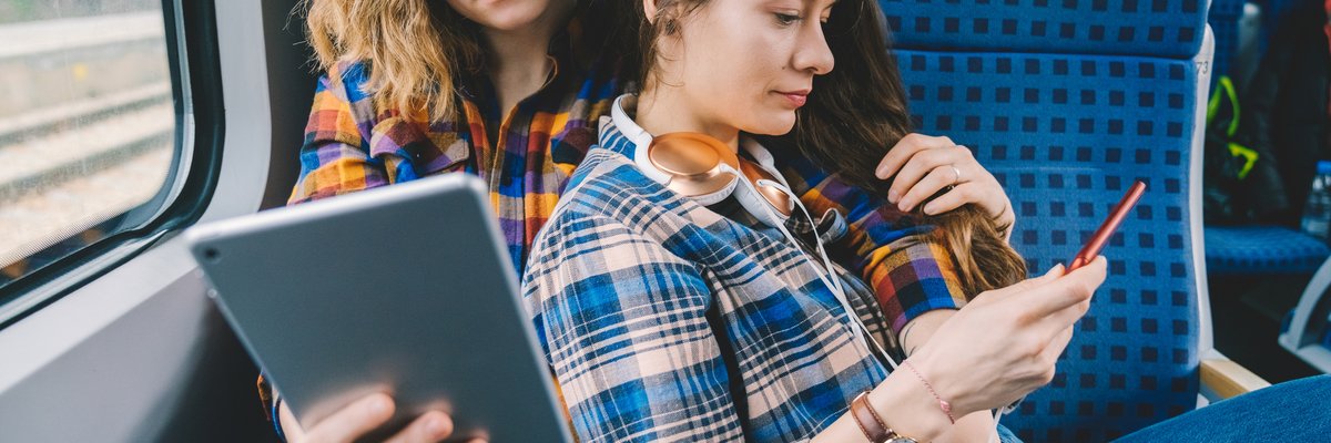 A couple sit together on a train. One looks at her tablet while the other looks at her phone.