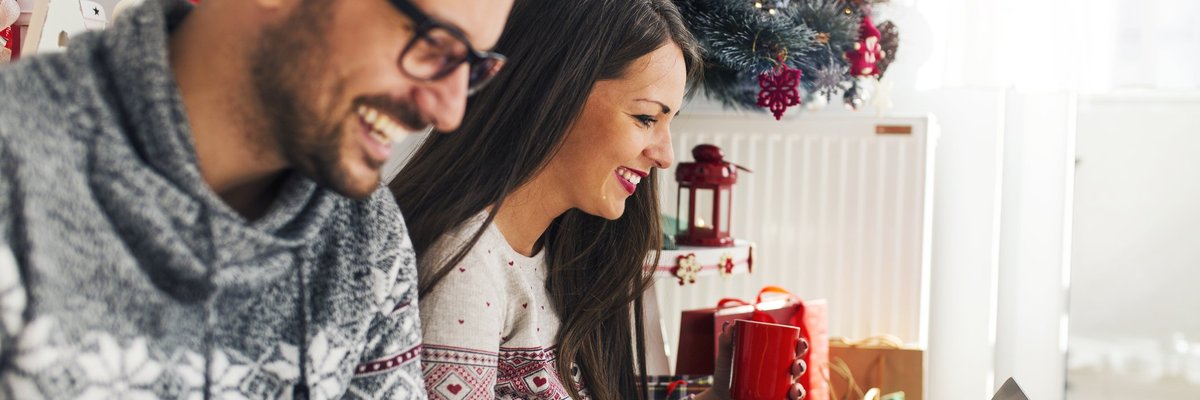 A smiling couple online shopping next to a Christmas tree.