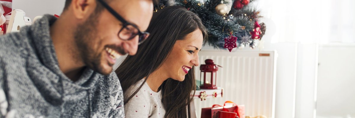 A smiling couple online shopping next to a Christmas tree.