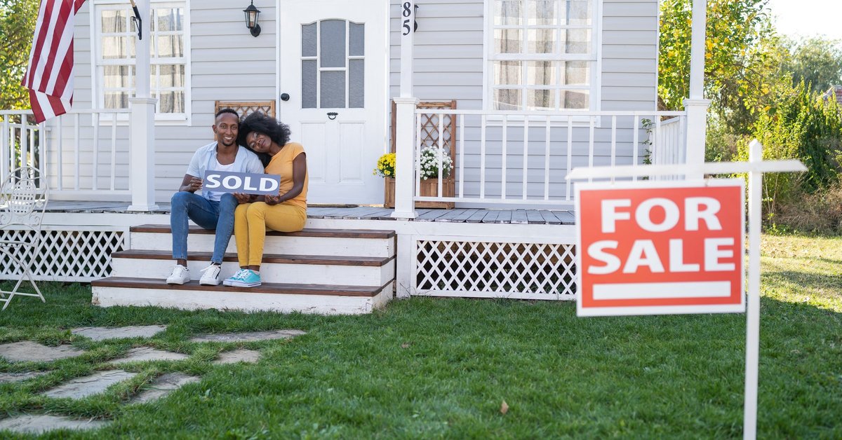 Couple sits on porch steps holding a house Sold sign.
