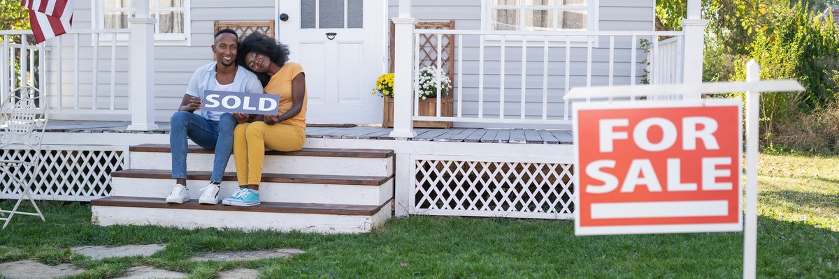 Couple sits on porch steps holding a house Sold sign.
