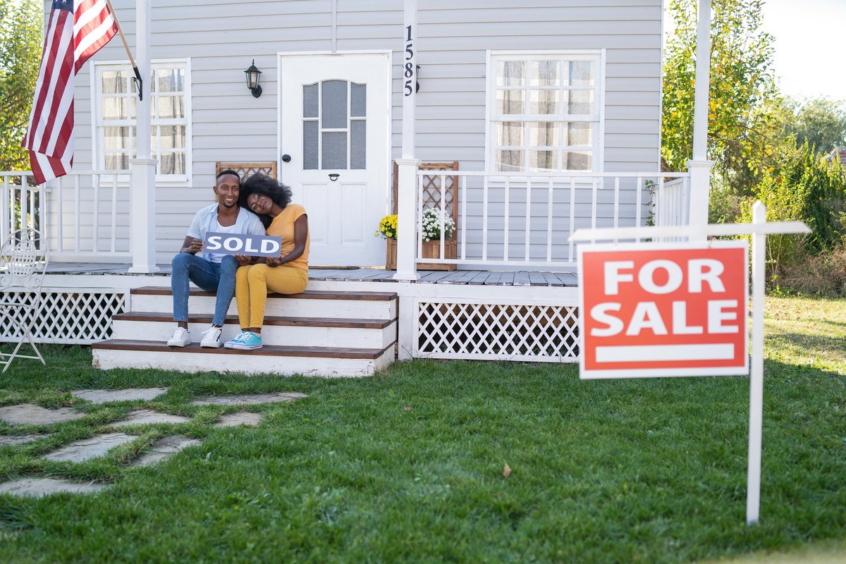 Couple sits on porch steps holding a house Sold sign.