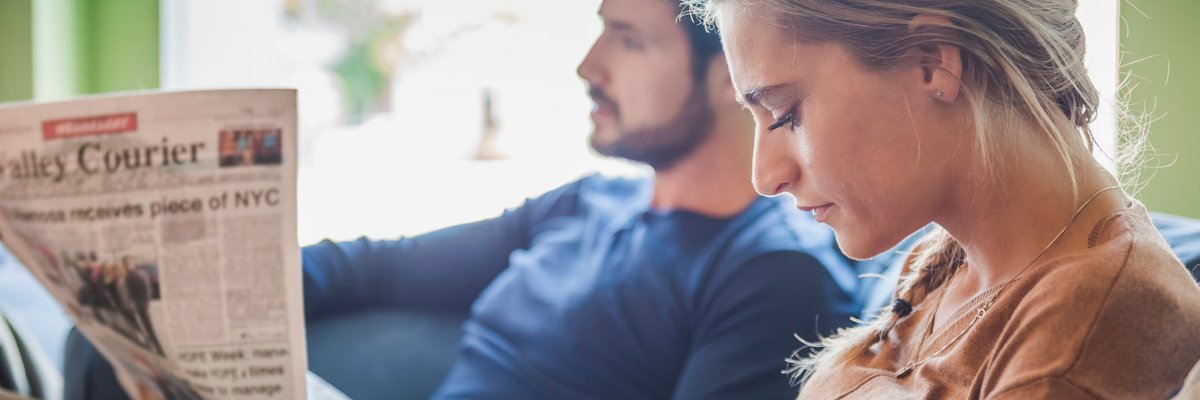 Couple sits together while one reads a newspaper and the other browses social media