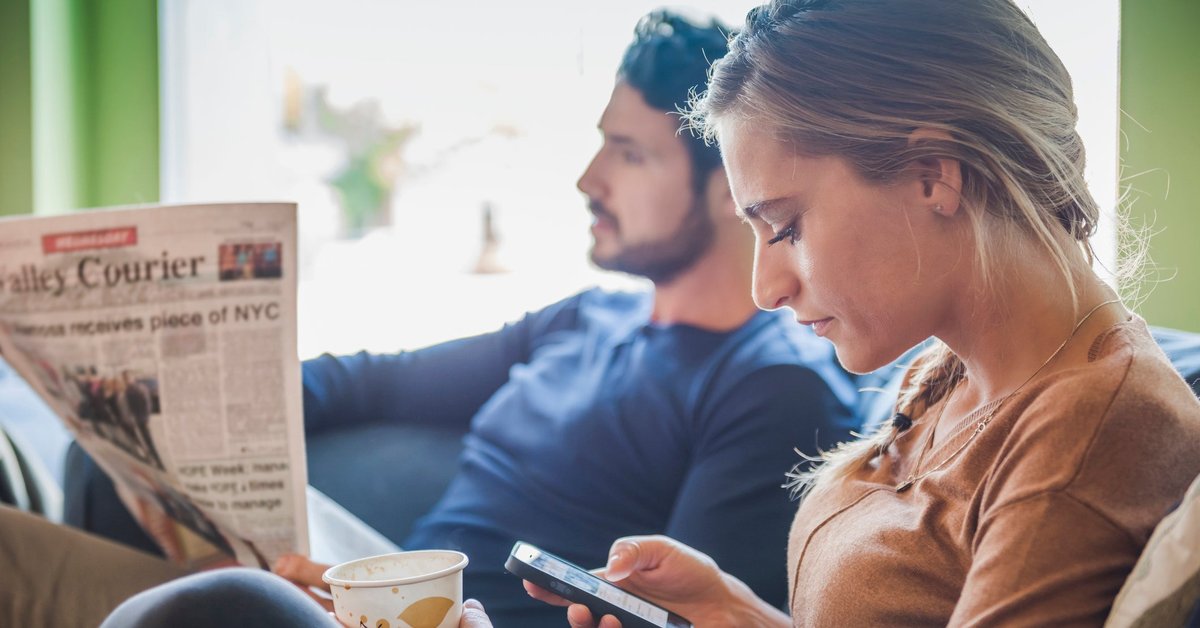 Couple sits together while one reads a newspaper and the other browses social media