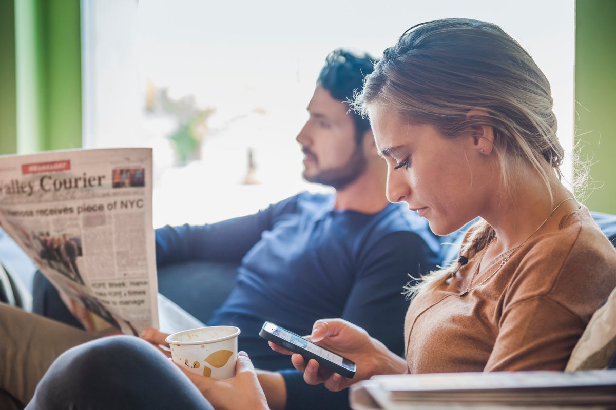 Couple sits together while one reads a newspaper and the other browses social media