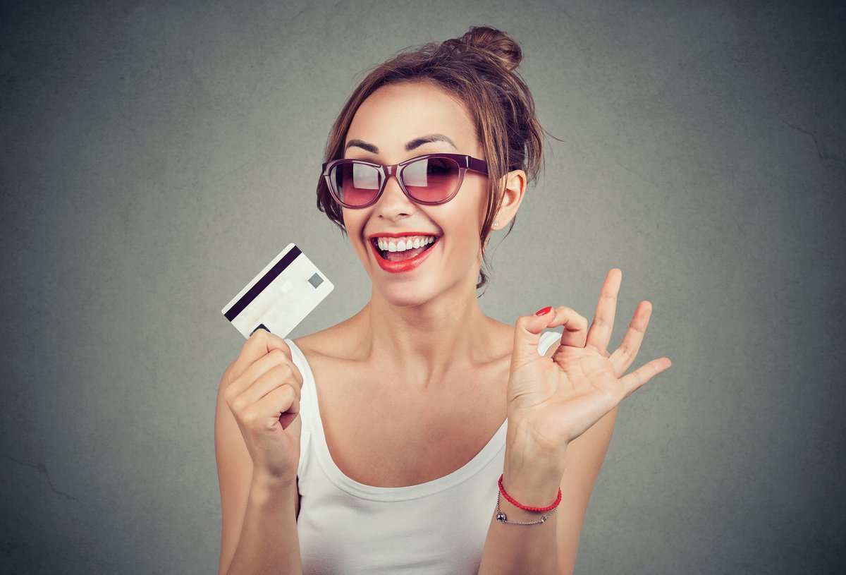 Woman wearing sunglasses holds a credit card in one hand and makes the OK sign with the other.