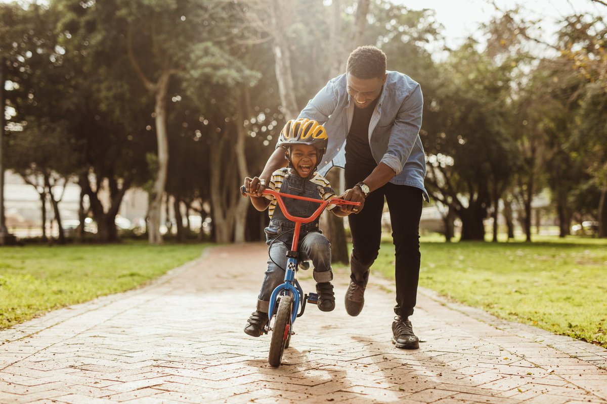 A dad helping his laughing young son ride a bike through a park.