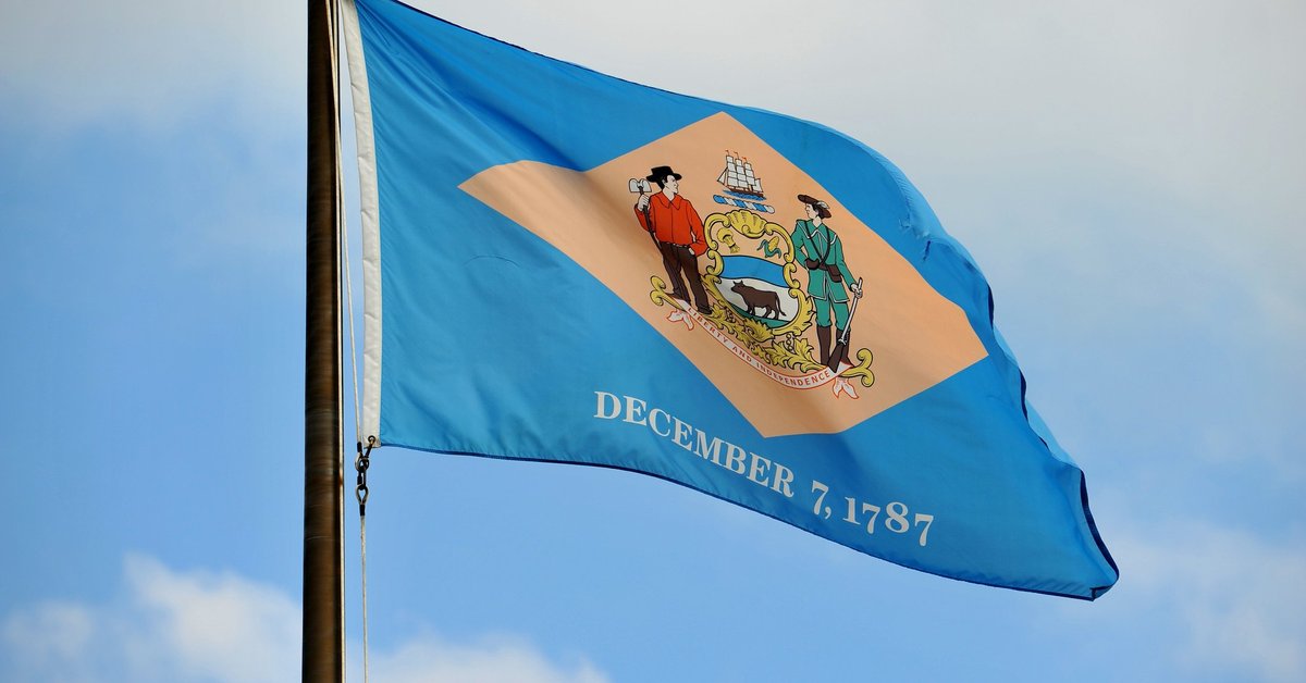 The Delaware state flag flying in front of a blue sky and white clouds.
