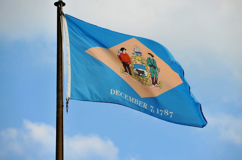 The Delaware state flag flying in front of a blue sky and white clouds.
