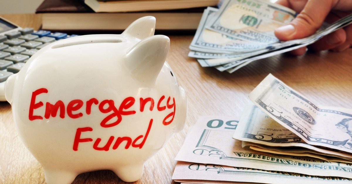 A white piggy bank that says emergency fund next to a stack of U.S. currency.