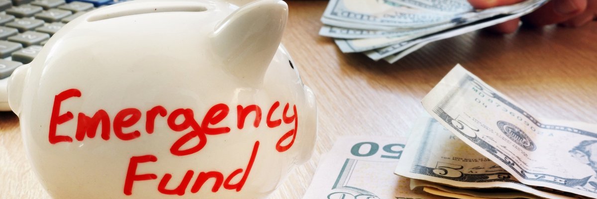 A white piggy bank that says emergency fund next to a stack of U.S. currency.