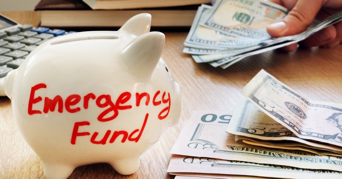 A white piggy bank that says emergency fund next to a stack of U.S. currency.