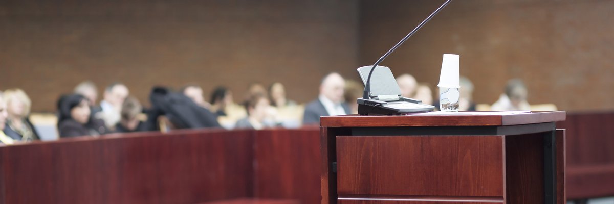 Empty mic stand in a courtroom with people seated in the background.